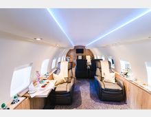 Innenraum eines luxuriösen Privatjets mit Ledersitzen, kleinen Esstischen, Holzverkleidung und Beleuchtung in sanftem Blau. An den Seiten befinden sich mehrere Fenster, und auf den Tischen sind Getränke und Snacks arrangiert.