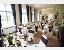 Ein festlich dekorierter Veranstaltungssaal mit weißen Tischdecken und Stuhlhussen, die mit braunen Schleifen verziert sind. Große Fenster mit dunklen Vorhängen und Kronleuchter verleihen dem Raum ein elegantes Ambiente; die Tische sind mit Blumen und goldenen Servietten dekoriert.