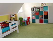 Kinderzimmer mit grünem Teppichboden. Links steht ein weißes Kinderbett, rechts ein Regal mit bunten Aufbewahrungsboxen, in der Mitte ein kleiner Tisch mit bunten Stühlen. Im Hintergrund ist eine Dachschräge und ein Fenster zu sehen.