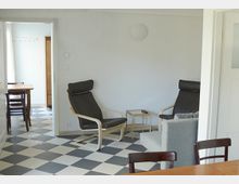 Wohnzimmer mit hellem Wänden und einem schwarz-weiß karierten Fliesenboden. Zwei gepolsterte Stühle mit Holzrahmen stehen neben einem kleinen Beistelltisch, im Hintergrund ist ein Durchgang zu einem angrenzenden Raum mit Esstisch und weiteren Stühlen erkennbar.