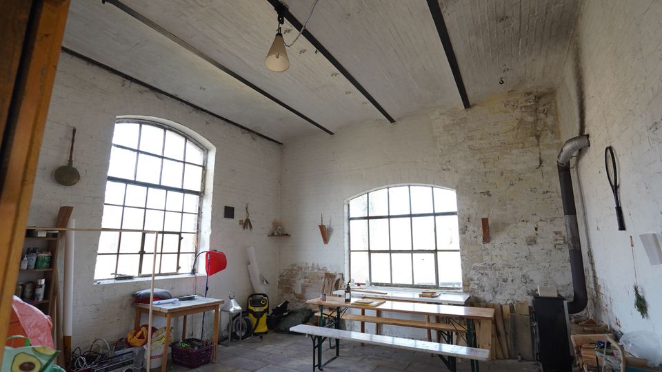 Innenraum einer rustikalen Werkstatt in einem alten Backsteingebäude mit weiß gestrichenen Wänden, Tonnengewölbe und freiliegenden Deckenbalken. Zwei große, segmentbogige Sprossenfenster, lange Holzbänke und Tische sowie ein schwarzer Ofen mit Rauchrohr stehen zwischen Werkzeugen, Holzstapeln und gelagerten Materialien.