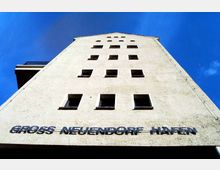 Blick auf die Fassade eines mehrstöckigen Gebäudes aus hellem Putz mit rechteckigen Fenstern in symmetrischer Anordnung und einem Schriftzug „Gross Neuendorf Hafen“ an der unteren Seite. Der Himmel ist klar und blau.