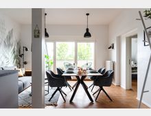 Moderner Ess- und Wohnbereich mit einem rechteckigen Esstisch und schwarzen Drehstühlen auf Parkettboden. Im Hintergrund große Fenster mit Blick ins Grüne, eine Balkontür, sowie eine graue Couch und eine Wanddekoration mit Pflanzenmotiv.