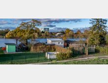 Kleingartenanlage mit mehreren kleinen Schuppen und Zäunen im Vordergrund, dahinter ein See mit Bäumen am Ufer. Im Hintergrund sind moderne Gebäude und eine halbrunde Konstruktion sichtbar, umgeben von herbstlicher Vegetation.