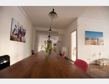 Langer Holztisch in einem hellen Raum mit weißen Wänden, roten und schwarzen Stühlen sowie hängenden Lampen. An den Wänden hängen Fotografien, und auf dem Tisch steht eine Vase mit Tulpen.