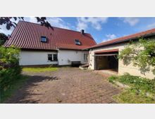 Einfamilienhaus mit rotem Satteldach, weißer Fassade und mehreren Fenstern, daneben eine offene Garage. Der Hof ist gepflastert und von etwas Grün umgeben, im Hintergrund leicht bewölkter Himmel.