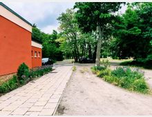 Seitenansicht eines orangefarbenen Gebäudes mit gepflastertem Gehweg und bepflanztem Beet entlang der Fassade. Daneben verläuft eine unbefestigte Zufahrt in einen baumbestandenen Grünbereich mit parkenden Autos und dichtem Laub.