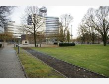 Grüne Parkanlage mit einem gepflasterten Gehweg und kahlen Bäumen im Vordergrund, im Hintergrund ein modernes Bürogebäude und der Funkturm von Berlin.