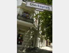 Straßenszene mit einem Altbau, dessen Fassade Fenster mit schwarzen Balkongeländern und Blumenkästen schmücken, sowie einem Straßenschild mit der Aufschrift „Gierkeplatz“. Im Vordergrund sind Pflastersteine und ein Bürgersteig mit angrenzenden Bäumen zu sehen, die Schatten auf die Fassade werfen.