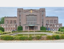 Backstein-Gebäude im neoklassizistischen Stil mit symmetrischer Fassade, großen hohen Fenstern und einem zentralen Eingang mit verglaster Tür und Vordach. Im Vordergrund ein rechteckiger Teich, umgeben von angelegten Grünflächen und Büschen, sowie ein Kopfsteinpflasterweg.