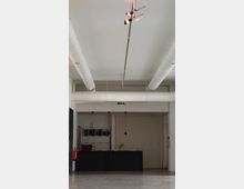 Ein minimalistischer Innenraum mit hellem Boden, weißer Decke und sichtbaren Lüftungsrohren. Im Hintergrund steht ein schwarzer Schreibtisch mit einer Pflanze, dahinter ein Regal und eine kleine Hängelampe.