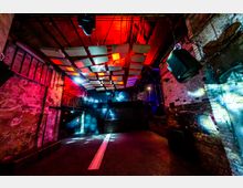 Innenraum eines Clubs mit industriellem Design, geprägt von freiliegenden Backsteinwänden, einer erhöhten Galerie und moderner Beleuchtung mit roten und blauen Lichteffekten. Die Decke ist mit akustischen Elementen in geometrischen Formen gestaltet, während Lautsprecher und Lichtinstallationen den Raum ergänzen.