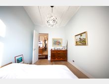 Schlafzimmer mit hellblauen Wänden, einer weißen Zimmerdecke mit Zierbalken und einem auffälligen, kugelförmigen Kronleuchter. Im Raum stehen ein antikes Holzsideboard mit Dekorationen und darüber hängen gerahmte Gemälde; im Hintergrund ist ein offener Durchgang zu einem hell erleuchteten Wohnbereich sichtbar.