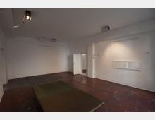 Ein leerer, weiß gestrichener Raum mit Betonboden in dunkler Farbe. Einfache Deckenspots beleuchten den Raum, der eine offene Tür, ein kleines rechteckiges Fenster und eine grüne Matte auf dem Boden hat.