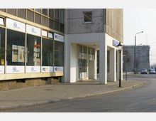 Außenansicht eines Reisebüros in einem modernen, mehrstöckigen Gebäude mit großen Glasfenstern und weißer Beschilderung. Im Vordergrund ist ein gepflasterter Gehweg, daneben eine Straße, im Hintergrund sind eine Straßenkreuzung und ein historisches Gebäude sichtbar.