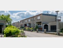 Reihenhäuser in einer Wohnsiedlung mit Flachdächern und verputzten Fassaden, umgeben von kleinen Vorgärten mit Büschen und Hecken; eine schmale Straße verläuft vor den Häusern.