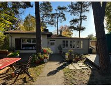 Eins-storyiges Haus mit Flachdach in einem Gartenbereich, umgeben von hohen Bäumen. Im Vordergrund befinden sich ein roter Holztisch, Gartenmöbel und Topfpflanzen auf einem gepflasterten Weg neben einer kleinen Holzterrasse.