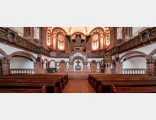 Innenraum einer Kirche mit roten Backsteinwänden, großen Rundbogenfenstern und einem hellen Gewölbedach. Im Vordergrund befinden sich Holzbänke, während der Altarbereich mit einer erhöhten Kanzel, einem Kruzifix und einer großen Orgel im Hintergrund dominiert wird.