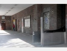 Überdachter Eingangsbereich im urbanen Stil mit roten Backsteinwänden, geschlossenen Rollläden an Fenstern, einer Tür mit Graffiti und einer eingebauten Uhr an der Wand. Rechts ist ein Eingang mit Rolltreppe zu sehen, und eine Person steht links in der Nähe der Tür.