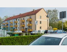 Mehrfamilienhaus mit vier Stockwerken in hellgelber Fassade und orangefarbenen Balkonen, umgeben von geparkten Autos, einer niedrigen Hecke und Straßenschildern. Im Hintergrund sind Bäume und Büsche zu sehen.