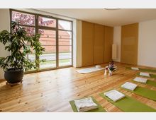 Heller Yoga- oder Meditationsraum mit Holzboden, großen Fenstern und Blick auf eine rote Backsteinmauer. Auf dem Boden liegen grüne Yogamatten mit gefalteten Decken, daneben eine Zimmerpflanze, Kerzen und ein Blumenarrangement.