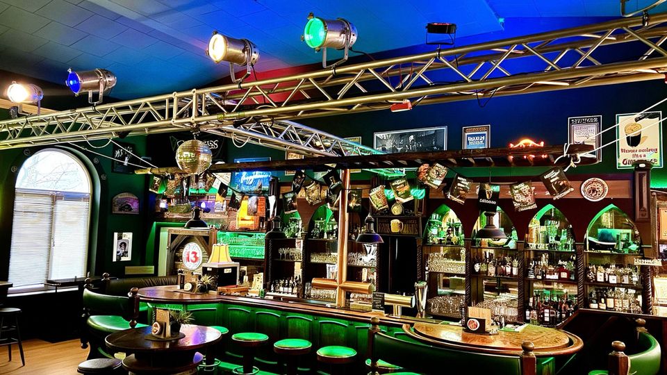 Innenraum einer Bar mit langem Tresen, grüner Beleuchtung und gepolsterten Barhockern sowie runden Tischen im Vordergrund. Über dem Tresen hängt eine Traverse mit bunten Scheinwerfern und einer Discokugel; im Hintergrund stehen beleuchtete Regale mit Flaschen und Gläsern sowie Wanddekorationen.