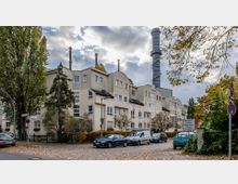 Mehrstöckiges Gebäude mit heller Fassade, flach geneigten Dächern und markantem Schornstein im Hintergrund. Im Vordergrund befinden sich eine gepflasterte Straße mit geparkten Autos, Bäume und ein Verkehrsschild.