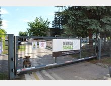 Ein Metalltor mit Schildern, darunter ein Warnhinweis und Geschwindigkeitsbegrenzung 10 km/h, verschließt den Eingang zu einem Firmengelände. Am Tor hängt ein Banner mit der Aufschrift „Demontage Schulz GmbH“, im Hintergrund sind Bäume, Gebäude und ein geparkter Transporter sichtbar; ein Hund sitzt hinter dem Tor.