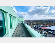 Außenbereich eines Gebäudes mit einer langen Terrasse, die mit grauen Bodenplatten und einer Metallbrüstung ausgestattet ist. Links befinden sich große Fensterfronten, rechts zeigt der Ausblick eine Stadtlandschaft mit Gebäuden, Straßen und Autos unter einem teilweise bewölkten Himmel.