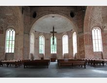 Innenraum einer Kirche mit freigelegtem Mauerwerk und hohen Rundbogenfenstern. Holzbänke sind in Reihen aufgestellt, ein schlichtes Holzkreuz hängt von der Decke, und im Hintergrund steht ein kleiner Altar.