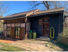 Einstöckiges Gebäude im rustikalen Stil mit einer Kombination aus verputzten Ziegelwänden und Holzfassade. Das Dach ist mit roten Ziegeln gedeckt, und vor dem Gebäude befinden sich ein gepflasterter Weg, ein Wasserschlauch an der Wand und zwei grüne Regentonnen.