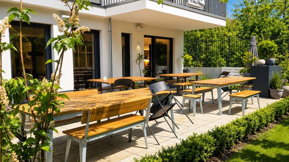 Gepflasterte Außenterrasse vor einem modernen, weißen Gebäude mit großen Glasfenstern und Balkon darüber. Mehrere lange Holztische mit Bänken und Klappstühlen stehen zwischen Hecken, Pflanzkübeln und dichtem Grün hinter einem Metallzaun.