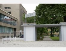 Eingang zu einem modernen Gebäude mit grauer Fassade, großen Fenstern und danebenliegendem Tor. Im Hintergrund sind ein kleiner Park mit Bäumen und ein gepflasterter Weg zu sehen.