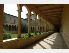 Überdachter Säulengang mit Rundbögen, flankiert von gelben Backsteinmauern und verzierten Säulen, der zu einem begrünten Innenhof mit umgebenden Gebäuden führt. Das Holzdeck der Decke und rankende Pflanzen auf der Mauer verleihen dem Gang Struktur und Atmosphäre.