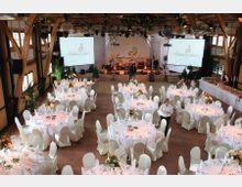 Großer Veranstaltungssaal mit Holzbalkenkonstruktion, dekoriert für eine Feier mit runden Tischen, die weiß eingedeckt und mit Blumenarrangements versehen sind. Im Hintergrund befindet sich eine Bühne mit Musikinstrumenten und zwei großen Leinwänden.
