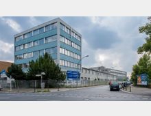 Mehrstöckiges Bürogebäude mit blau-grauer Fassade und großen Fenstern in einer städtischen Umgebung. Im Vordergrund ein Zaun, einige Bäume, parkende Autos entlang einer Straße sowie eine Werbetafel; bewölkter Himmel im Hintergrund.