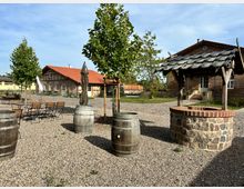 Ein ländlicher Innenhof mit Kiesboden, Holztischen und -stühlen, hölzernen Weinfässern und einem gemauerten Brunnen mit Dach. Im Hintergrund sind zwei Backsteingebäude mit roten Ziegeldächern und grüner Umgebung zu sehen.