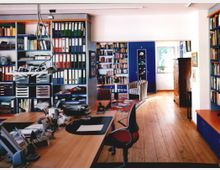 Ein Büro mit Holzboden, einem großen Schreibtisch im Vordergrund und Wandregalen voller Ordner und Bücher. Im Hintergrund befindet sich ein Sitzbereich mit Sesseln, einem Regal und einem Fenster, das Tageslicht hereinlässt.