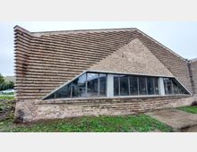 Ein modernes Backsteingebäude mit einer markanten, gestuften Fassadengestaltung aus vorspringenden Steinlagen. Ein großes, trapezförmiges Fenster mit Metallrahmen ermöglicht Einblick in den Innenraum; der umgebende Bereich ist mit Rasen und Gehwegplatten versehen.