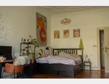 Bright bedroom with high ceilings, white walls, and a black platform bed with striped bedding, surrounded by framed art and a large colorful wall tapestry. A small desk and shelving unit with plants sit by the bed, and an open doorway leads to an adjacent room over wooden floors.