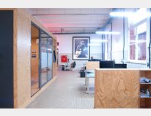 Ein modernes Büro mit einem offenen Raumkonzept, das Holzmöbel und Arbeitsbereiche mit Computerbildschirmen zeigt. Große Fenster auf der rechten Seite lassen Tageslicht herein, während die Wände mit weißen Fliesen abgesetzt sind; im Hintergrund sind eine Glaskabine und ein großes Wandbild zu sehen.