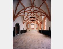 Gotisches Kircheninterieur mit hoch aufragenden roten Kreuzrippengewölben, großen spitzbogenförmigen Fenstern und schmiedeeisernen Kronleuchtern. Der Raum hat ein Steinpflaster und dunkle Holzvertäfelungen an den Seitenwänden.