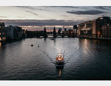 Blick auf die Spree bei Sonnenuntergang in Berlin, mit der beleuchteten Oberbaumbrücke im Hintergrund und einem Boot im Vordergrund. Rechts ist ein modernes Gebäude mit dem Schriftzug "Universal" zu sehen, links alte und neue Fassaden entlang der Uferpromenade.