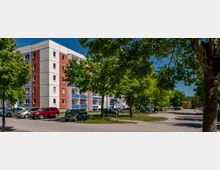 Mehrstöckiges Wohnhaus mit rot-weiß-grauer Fassade und Balkonen mit blauen Brüstungen, an einer ruhigen Straße. Vor dem Gebäude stehen geparkte Autos, und hohe Bäume säumen Gehweg und Grünstreifen.