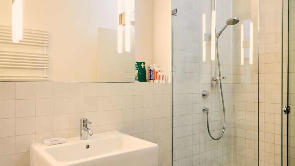 A modern bathroom with a white tiled wall, a glass-enclosed shower with a rainfall showerhead, and a wall-mounted rectangular sink with a mirror above it. The floor is dark-tiled, and a black trash bin is placed under the sink.