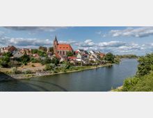 Stadtansicht mit einer Kirche im gotischen Stil und rotem Dach als zentralem Element, umgeben von Häusern mit bunten Dächern, gelegen an einem Flussufer mit grüner Vegetation und klarem Himmel im Hintergrund.