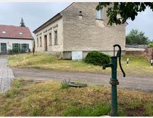 Eine ländliche Szene mit einer alten Handwasserpumpe im Vordergrund, umgeben von einer Wiese. Im Hintergrund stehen ein verputztes, teilweise renovierungsbedürftiges Gebäude und ein kleineres Haus mit roten Ziegeldächern und grünen Fensterläden.