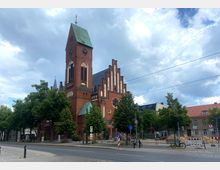 Rote Backsteinkirche mit einem hohen Kirchturm und grünem Spitzdach, umgeben von Bäumen, mit angrenzender Straßenkreuzung und Baustellenabsperrungen. Der Himmel ist bewölkt, und die Straße im Vordergrund zeigt Verkehrsschilder und ein Fahrrad.
