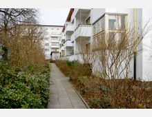 Ein Weg mit grauen Platten verläuft zwischen kahlen Sträuchern und einem modernen Wohngebäude mit weißen Fassaden und Balkonen auf der rechten Seite. Im Hintergrund sind weitere mehrstöckige Gebäude zu sehen.