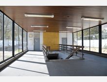 Innenraum eines einfachen Übergangsgebäudes mit großen Fenstern an den Seiten, die den Blick nach draußen erlauben. Der Raum hat eine Holzdecke, Neonleuchten, Treppengeländer aus Holz und Metall, sowie eine Treppe, die nach unten führt; im Hintergrund eine Tür, eine Wanduhr und gelbe Wandverkleidung.